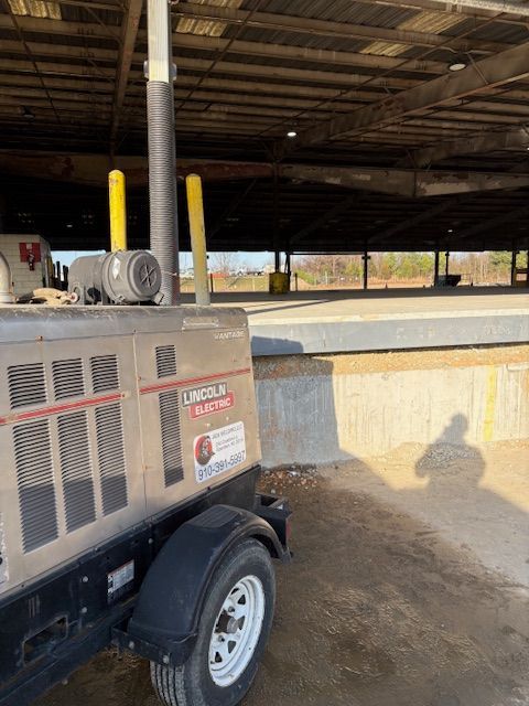 A tan Lincoln Electric towable welder parked on a dirt lot under a large, open-sided covered structure.