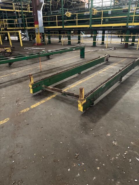 Two parallel green industrial chain conveyor tracks sit on a concrete warehouse floor beneath an elevated metal platform.