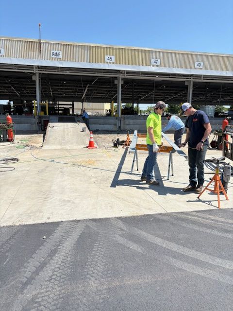 Two workers in high-visibility and work clothing stand at a metal workbench outdoors, preparing materials for construction.