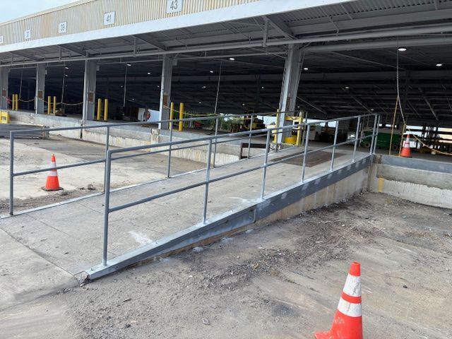 A concrete ramp with metal railings leading into an open-air industrial warehouse, marked by two orange safety cones.