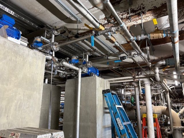 Industrial utility room ceiling showing blue electric pumps, network of metal pipes, and blue ladders below.