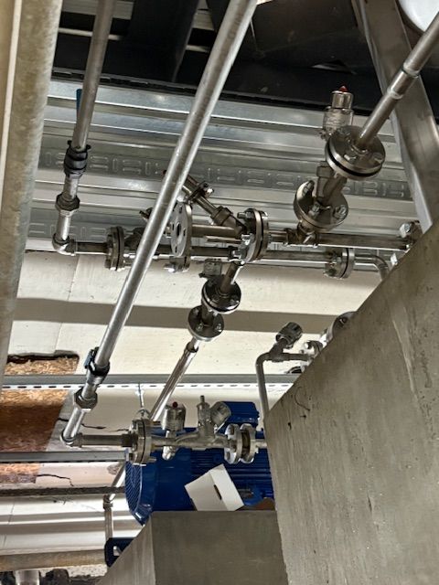 Stainless steel piping and flanges mounted on a ceiling above a blue motor in an industrial setting.