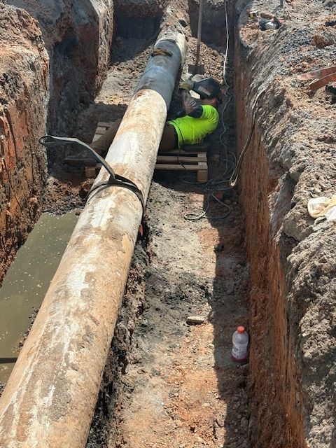 A worker in a high-visibility vest sits inside a deep, narrow trench beside a large pipe.