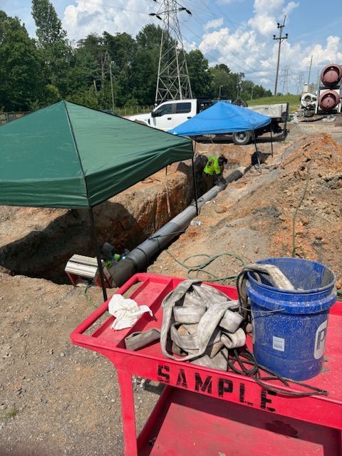 A construction site with an open trench, a long pipe, two tents, and a red utility cart in the foreground.