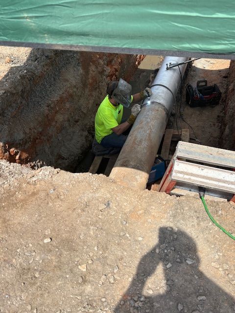 A worker in a bright yellow shirt and welding helmet repairs a large metal pipe inside an earthen trench.