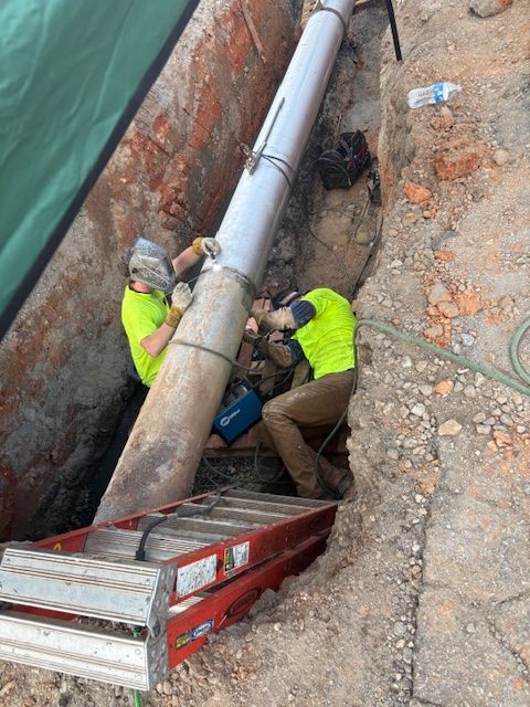 Two construction workers in neon yellow vests weld a metal pipe together inside a dirt trench.
