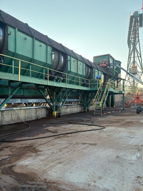 A large, green industrial aggregate or hopper system stands outdoors on a concrete ground, with a tall crane in the back.