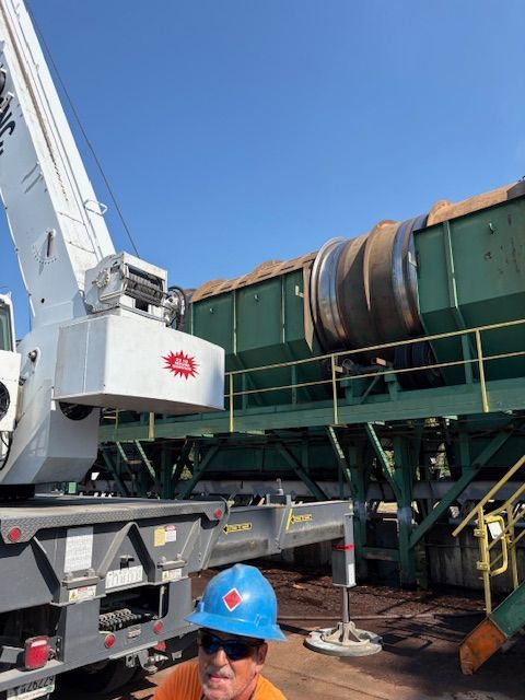 A crane positioned next to a large industrial green machine containing material, with a worker in a blue hard hat nearby.