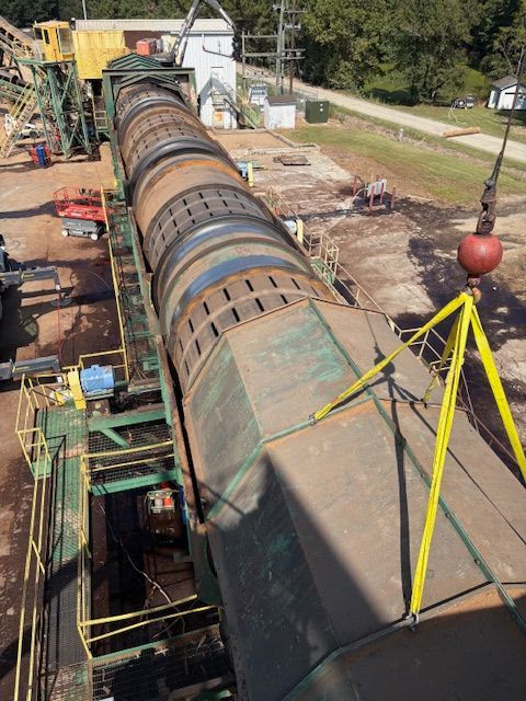 A large industrial rotary drum being lifted by a crane at an outdoor aggregate processing site.