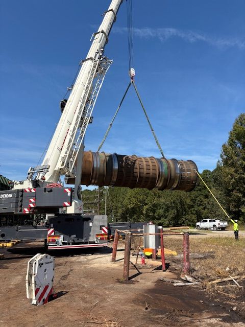 A crane lifts a large industrial cylindrical component at an outdoor site, with a worker nearby holding a guide rope.