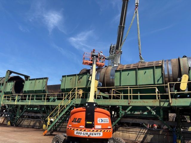 A construction crane lifts a large industrial cylindrical component onto a green frame while a worker operates a JLG lift.