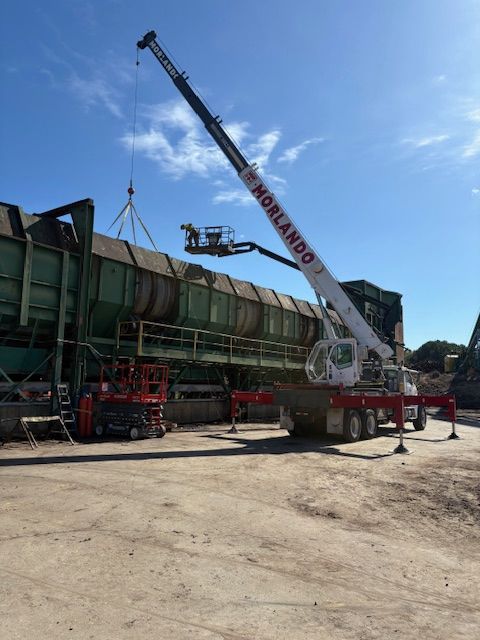 A white crane with the name Morlando works on a large green industrial structure under a blue sky.