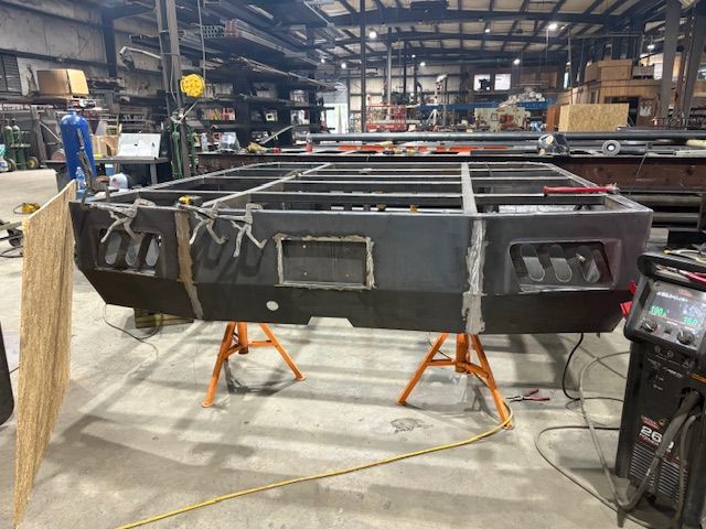 A large, grey metal frame for a vehicle bumper sits on orange jack stands inside an industrial welding workshop.