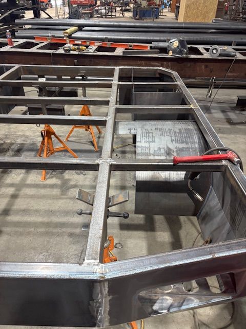 A metal framework for a vehicle trailer under construction in a workshop, with orange jack stands supporting the structure.