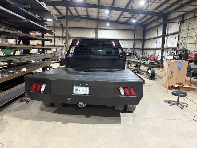 A black flatbed truck rear parked in a large metal fabrication workshop with shelves of metal stock and tools.