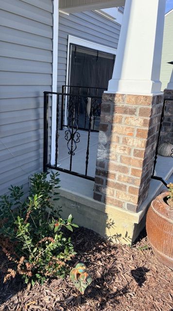 A side view of a porch with black wrought-iron railings, a tan brick pillar, light gray siding, and a small shrub.