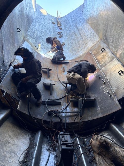 Three welders in protective gear work inside a large, cylindrical metal structure, sparks flying as they join metal plates.