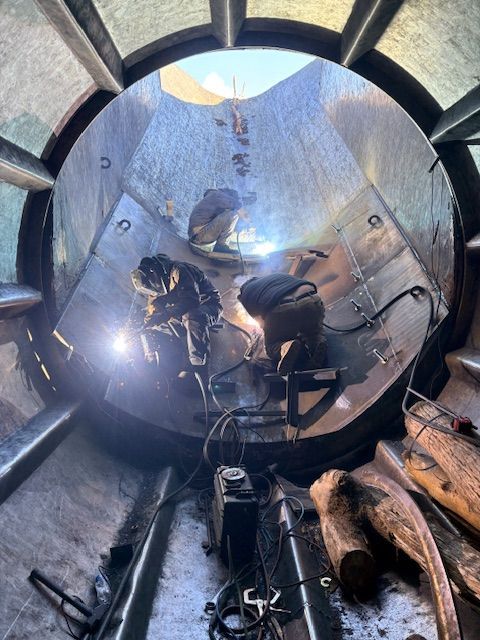 Two people in protective welding gear work on metal panels inside a large, cylindrical industrial structure.