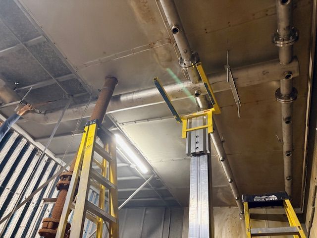 Two yellow ladders stand in an industrial ceiling space, one supporting a vertical metal beam against overhead piping.