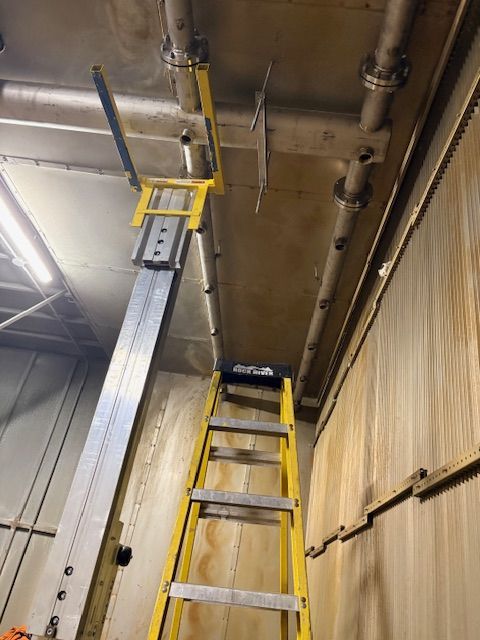 A yellow stepladder stands beneath a metal beam and ceiling pipes in an industrial setting.
