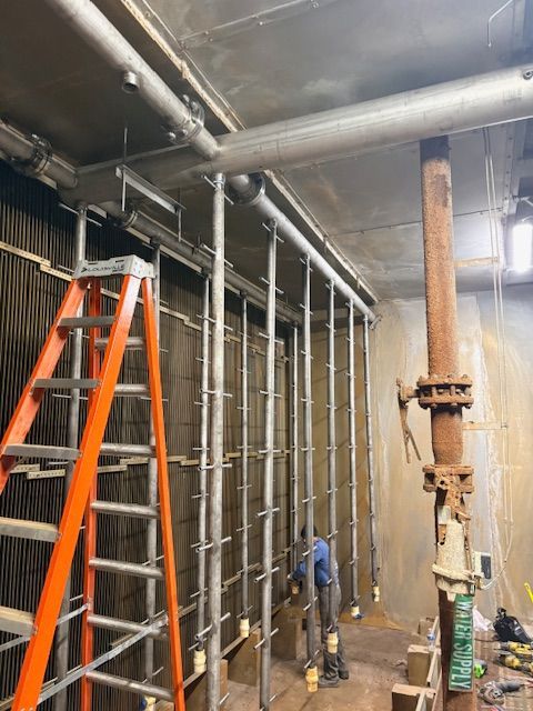 An orange ladder stands beside a worker installing vertical support poles in a construction site with exposed piping.