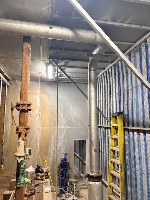 A vertical metal pipe and a rusted pipe with a valve stand in an industrial indoor space with a ladder and gas tank.