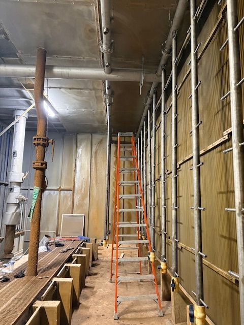 An orange stepladder stands in a narrow construction zone between a wall of wood panels and metal support poles.