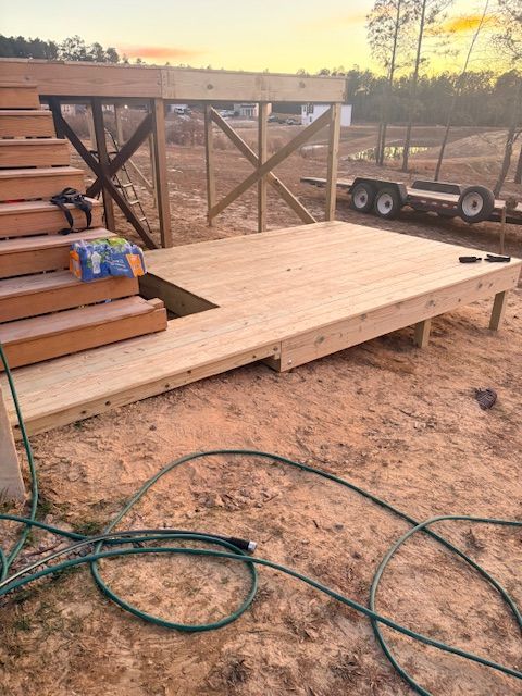 A wooden deck with a staircase under construction in an outdoor dirt lot with a utility trailer in the background.