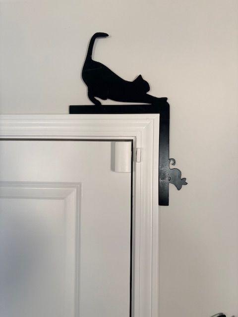 A black metal decorative topper shaped like a stretching cat sitting on the top corner of a white door frame.