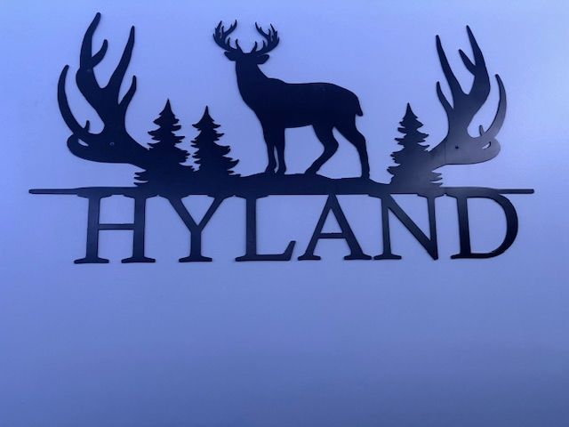 Black metal wall art featuring a deer silhouette, pine trees, and antlers, with the name 