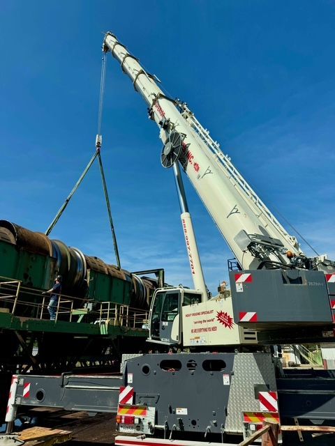 A large mobile crane lifting a green industrial cylindrical machine under a clear blue sky.