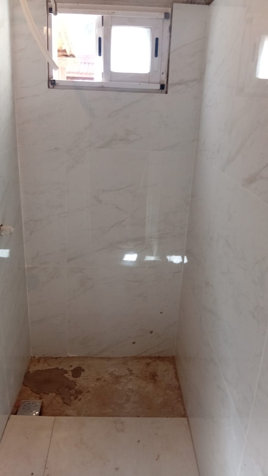 Baño estrecho con paredes de azulejos blancos y ventana. Suelo marrón.