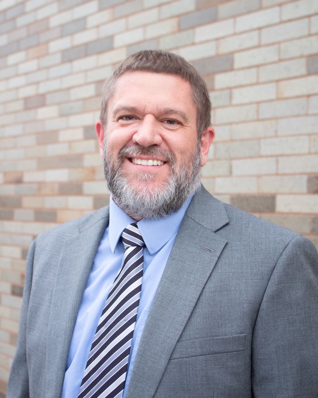 Man in grey suit smiles at camera, standing in front of a brick wall.
