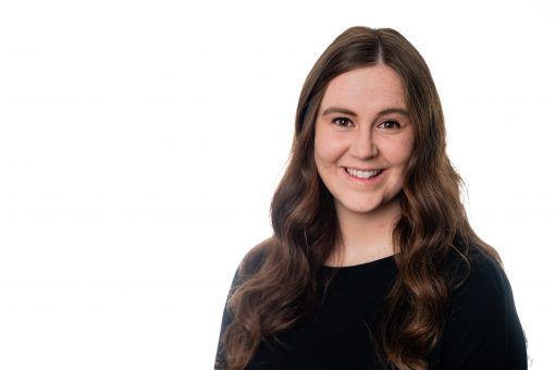 Woman with long, wavy brown hair, smiling at camera; wearing a black shirt against a white background.