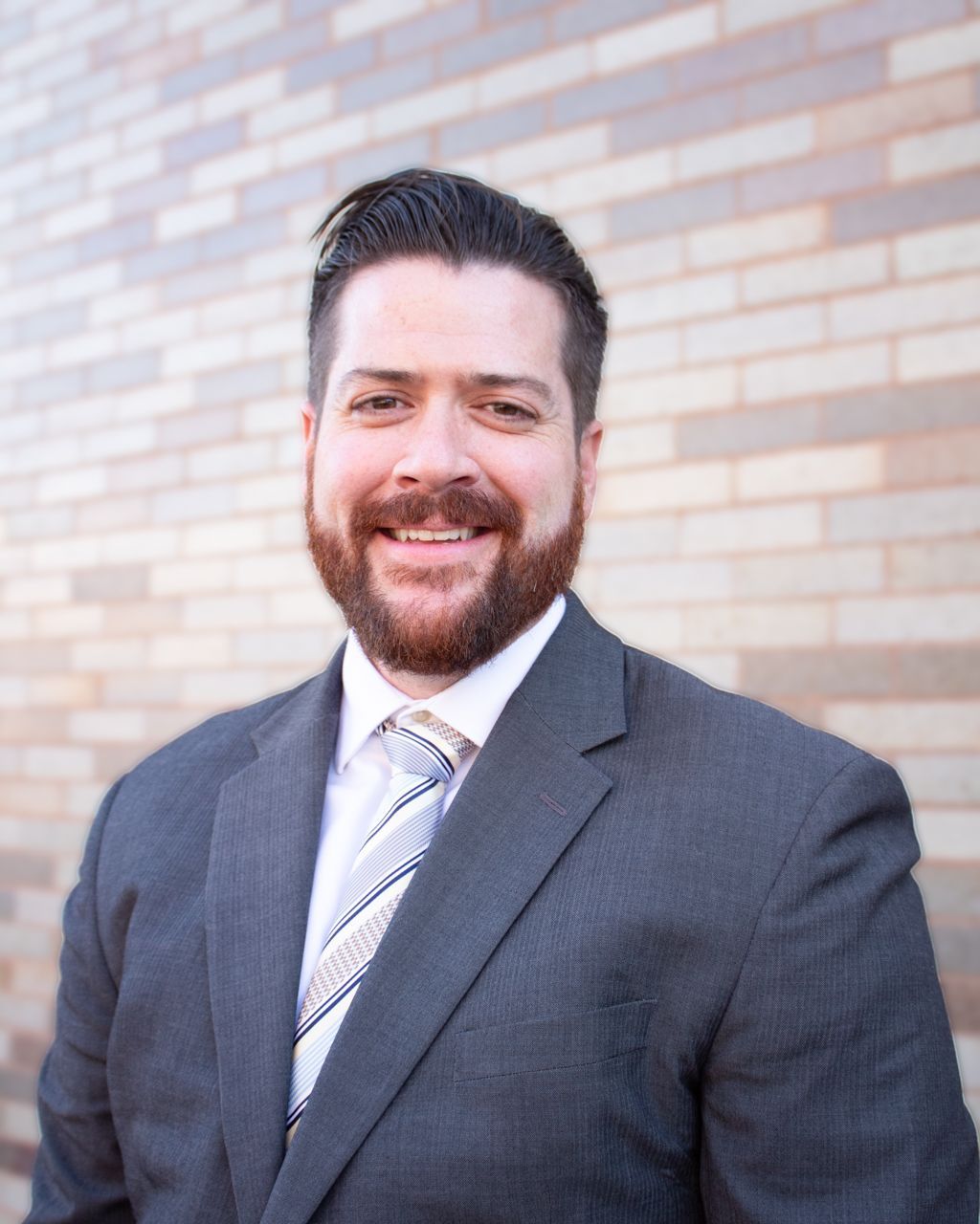 Man in gray suit with beard smiling in front of a brick wall.