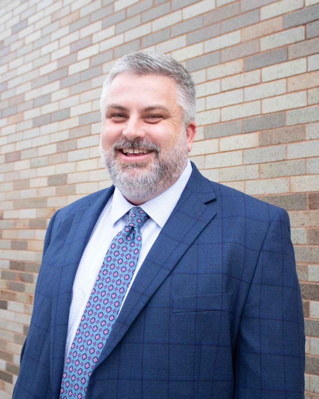 Man in blue suit, smiling, standing in front of a brick wall.