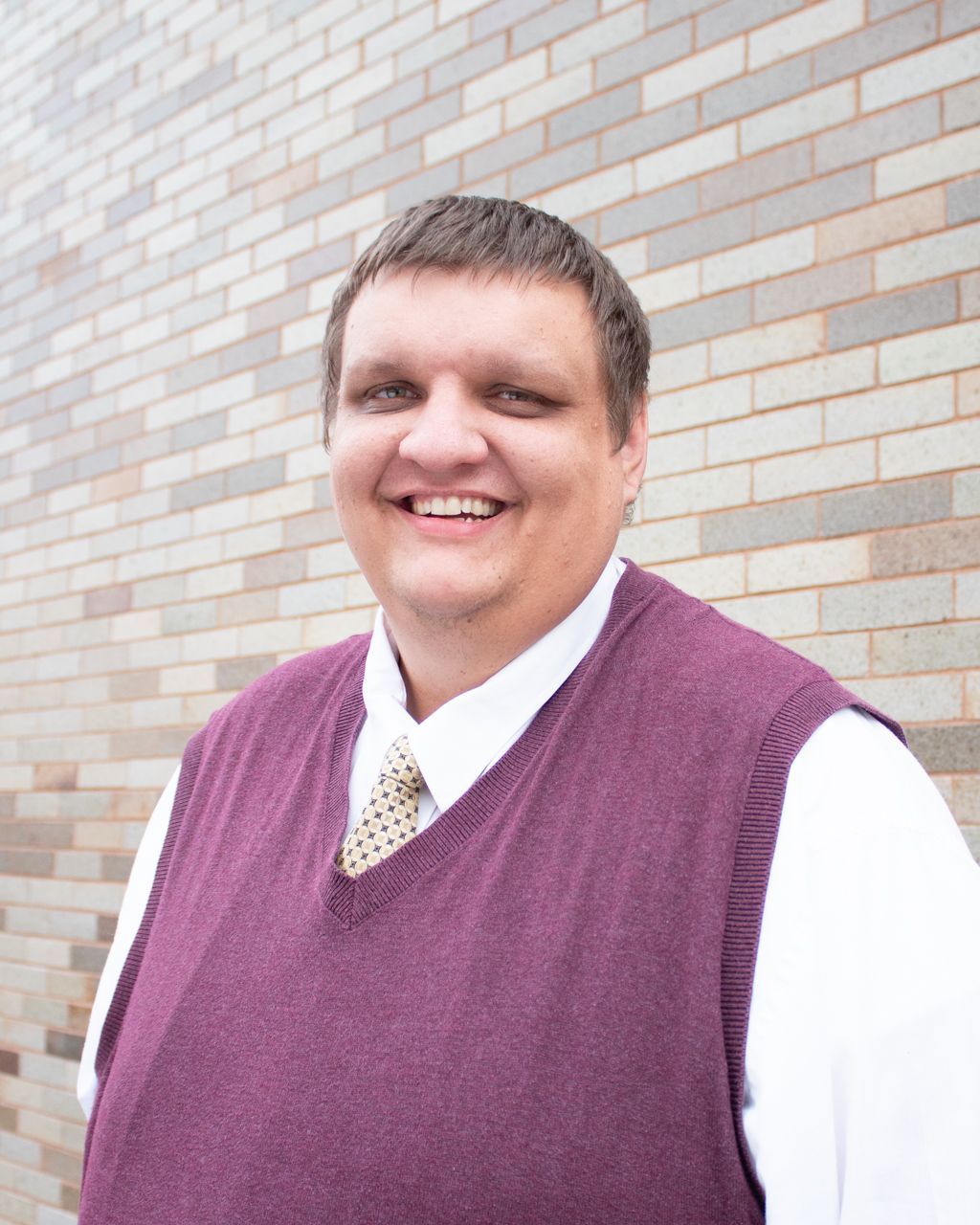 Man in a maroon sweater vest smiles in front of a brick wall. He wears a white shirt and patterned tie.