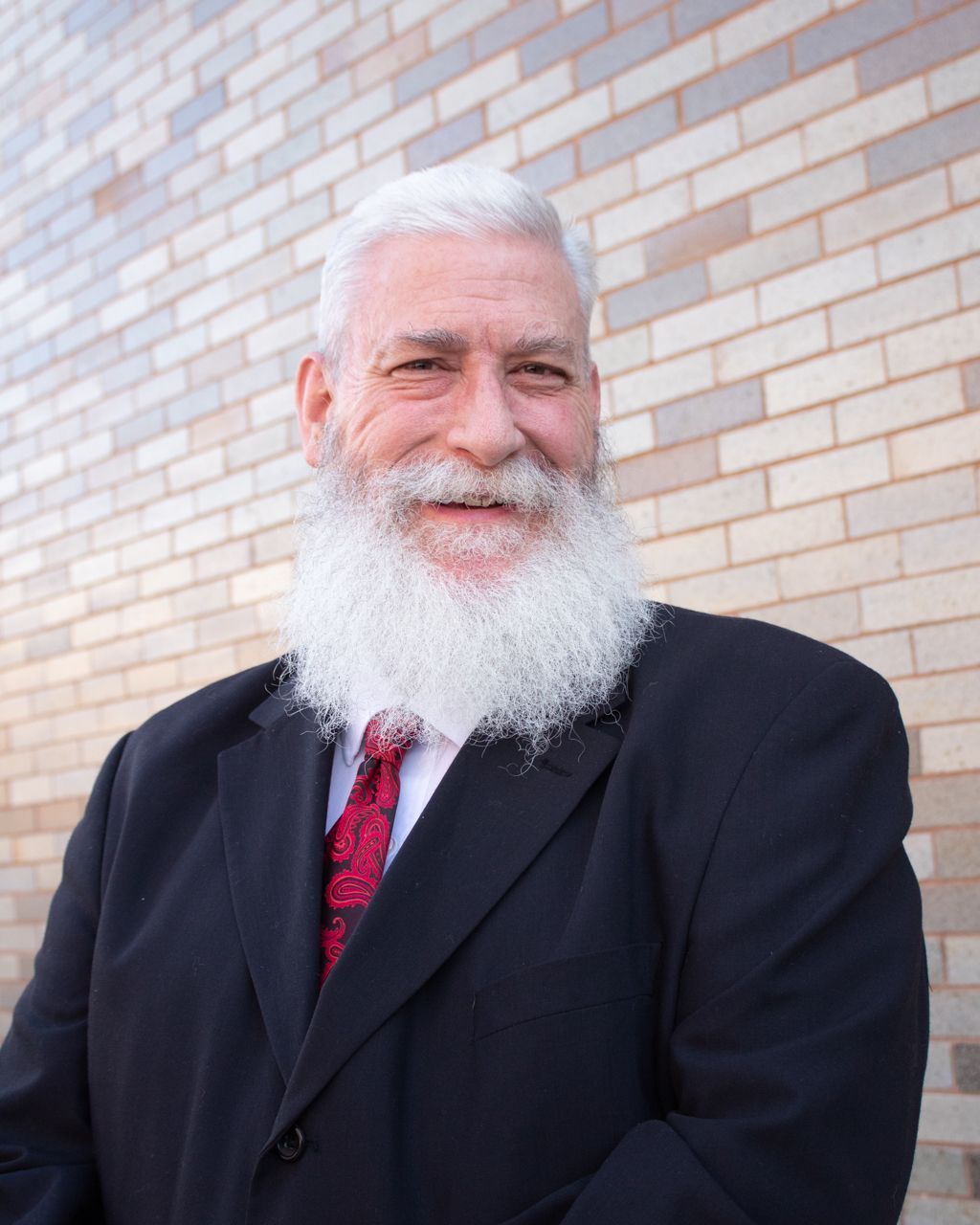 Man with a long, white beard wearing a black suit and red tie stands in front of a brick wall, smiling.