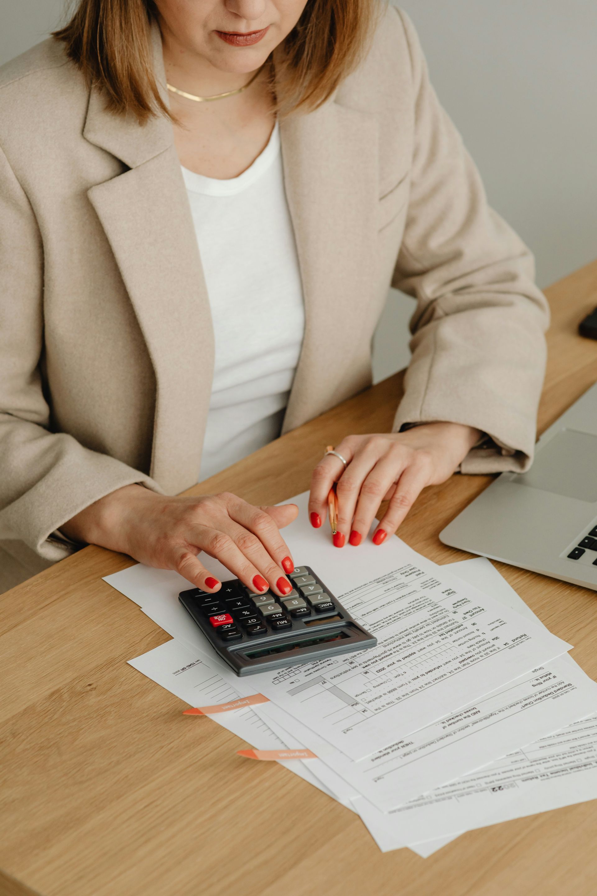 Woman in tan blazer using calculator, reviewing documents at a wooden desk.