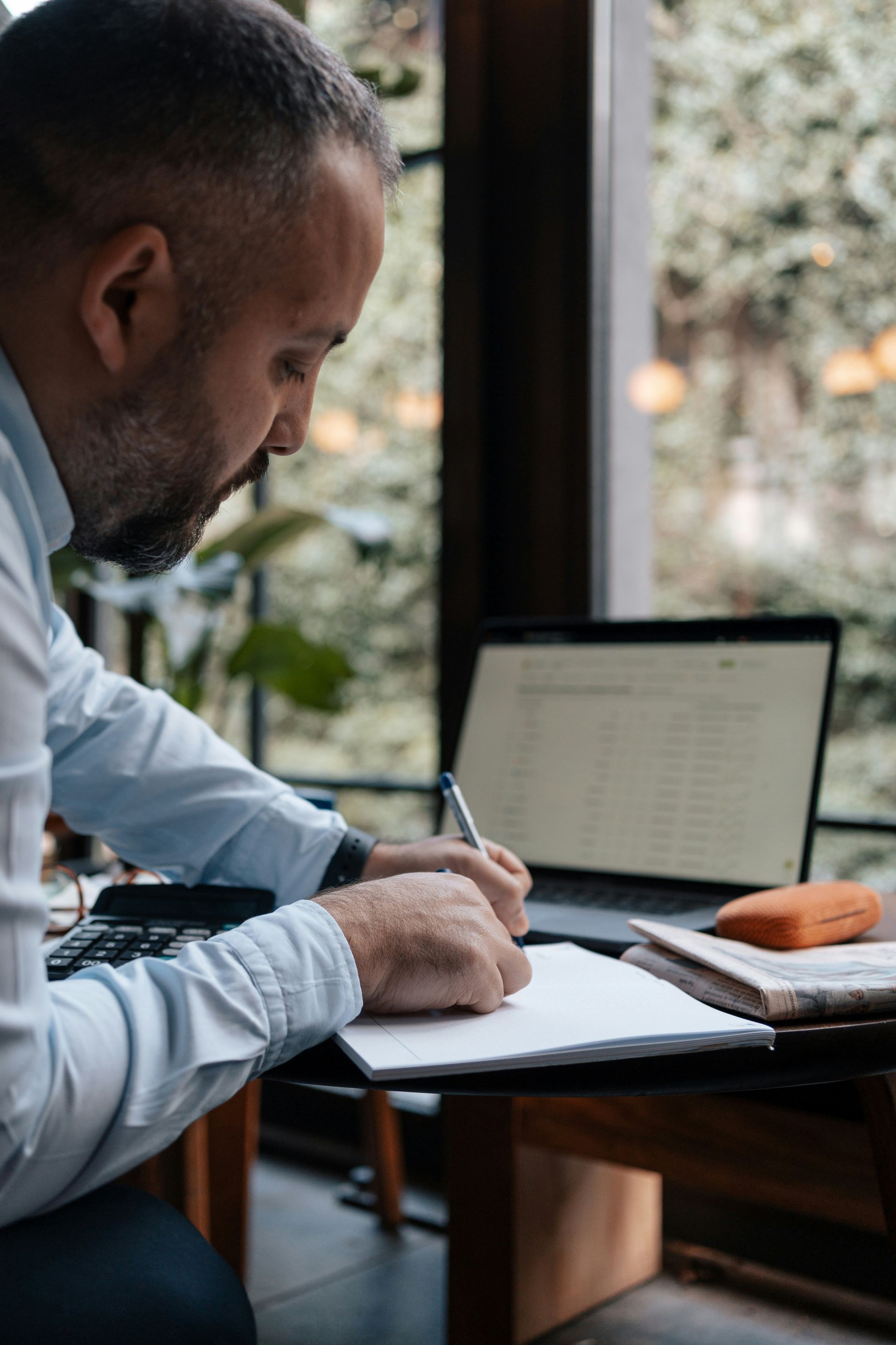 Man writing on paper, laptop in the background, near a window with greenery.