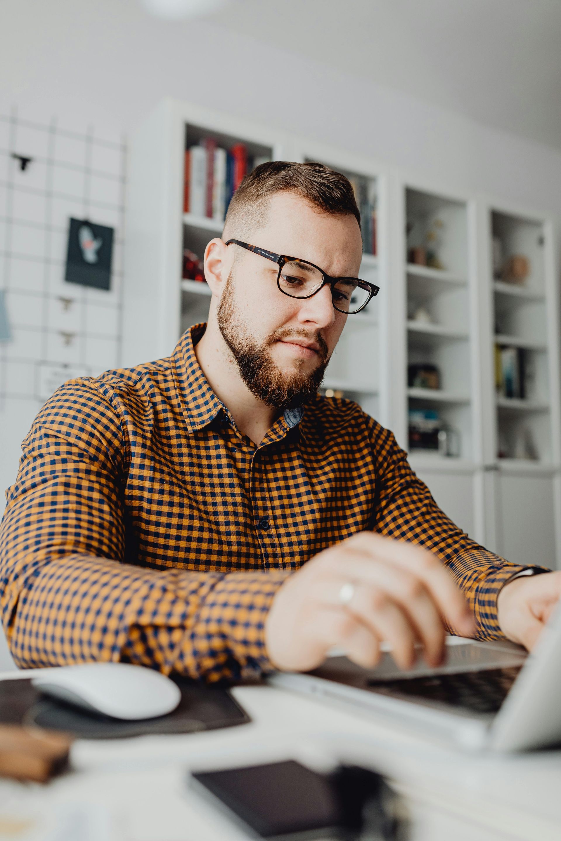 Man with glasses and beard, working on a laptop at a desk, with bookshelves in the background.