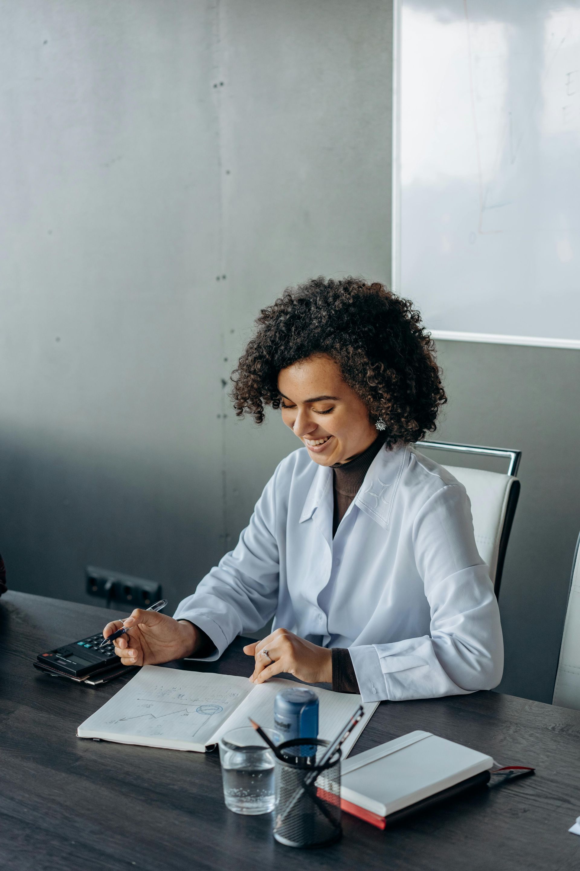 Woman in white coat, smiling, works at desk, using calculator and looking at papers.
