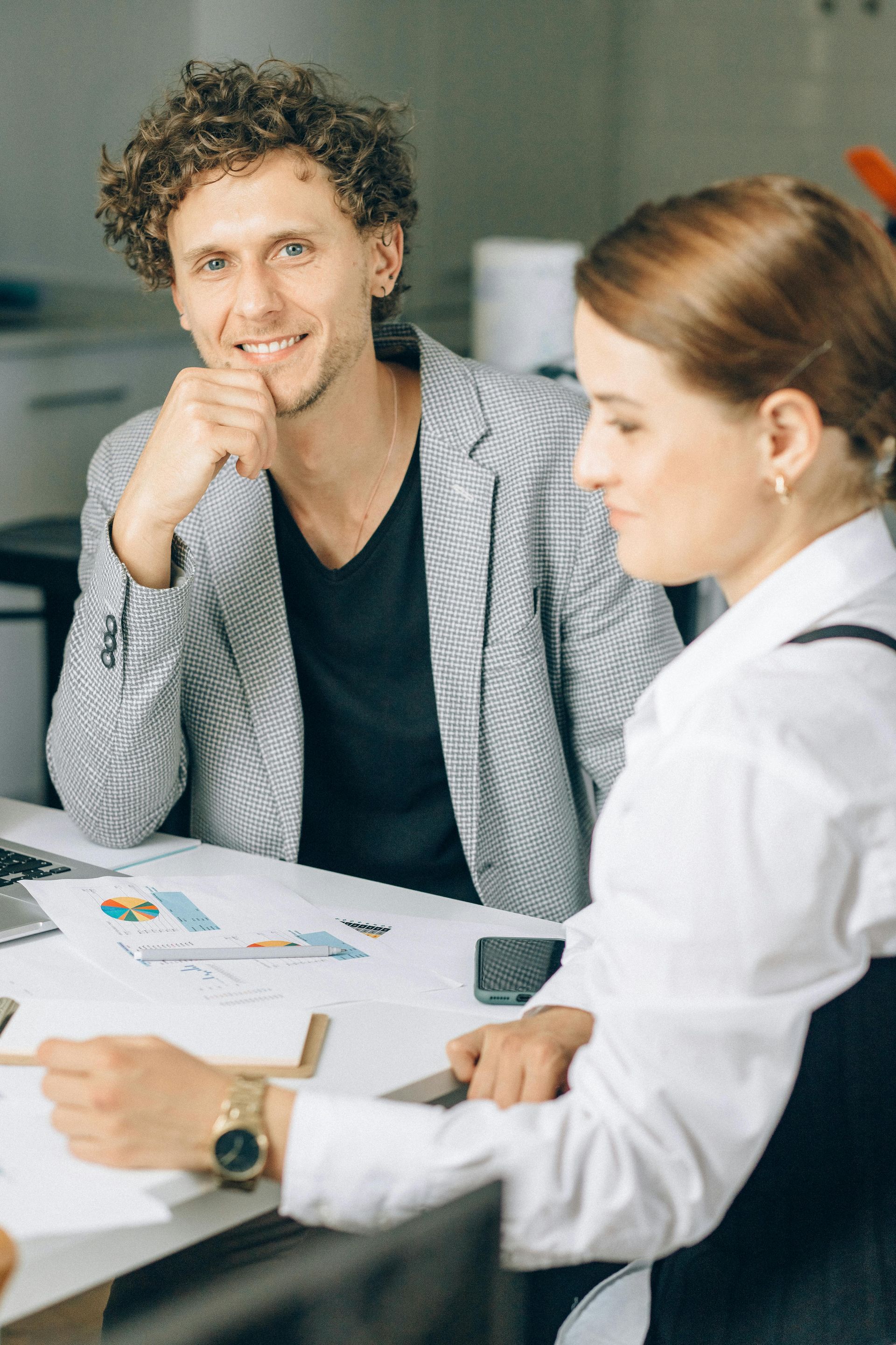 Man smiles, looking at the camera, while a woman beside him analyzes papers at a table.