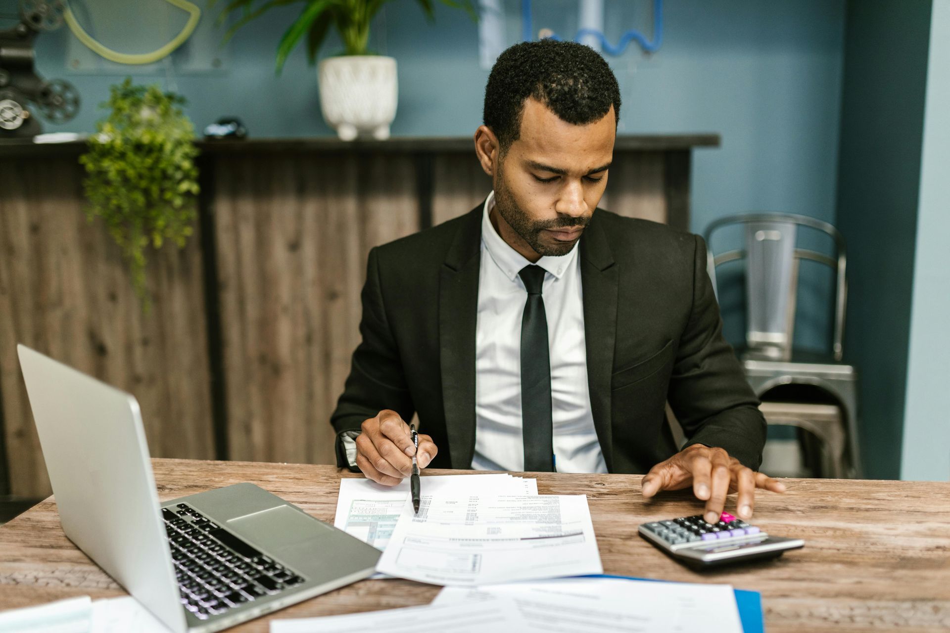 Man in suit working at a desk, using calculator and laptop, reviewing documents.