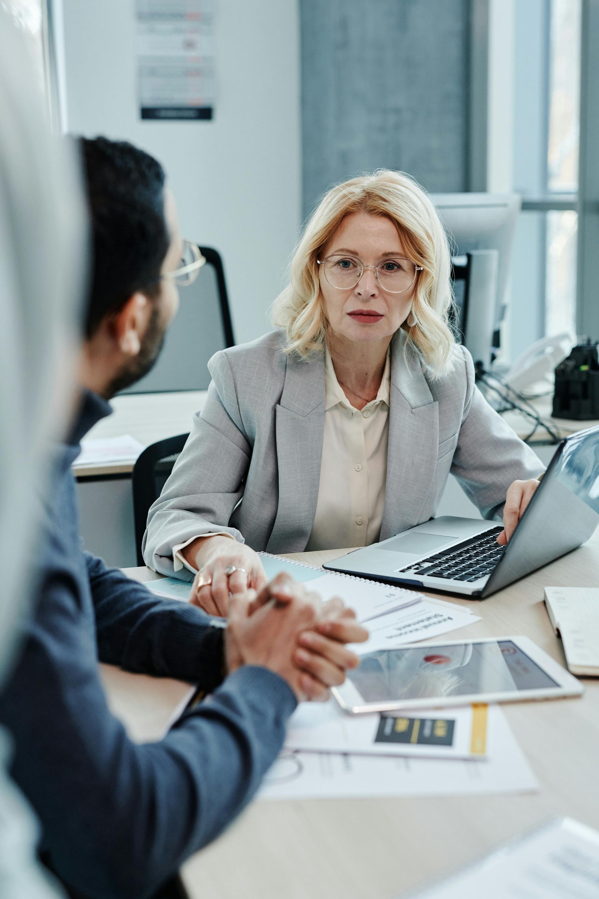 Woman with glasses and laptop gestures towards two people at a desk.