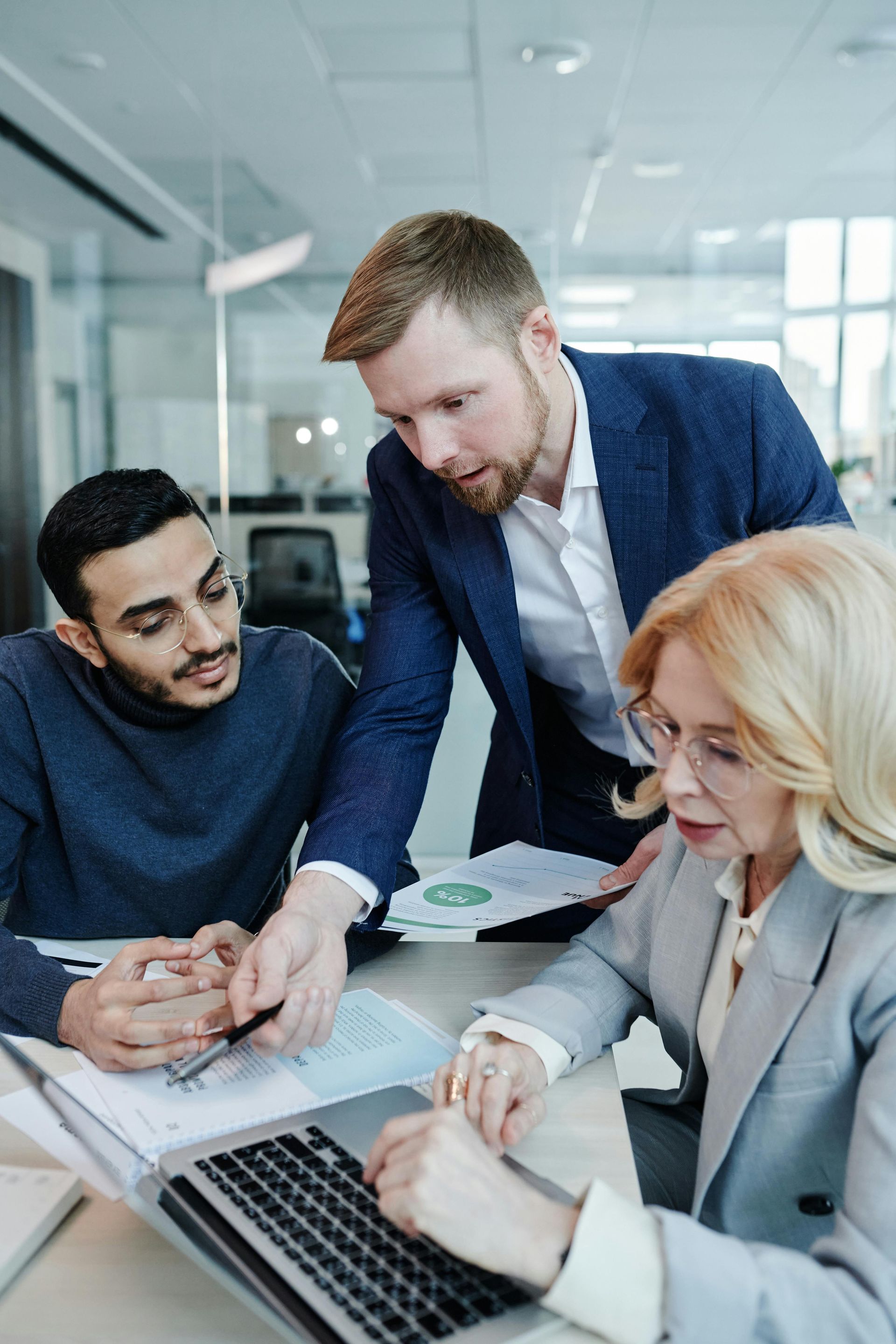 Three people collaborate at a table with papers and a laptop in a bright office. One points at a document.
