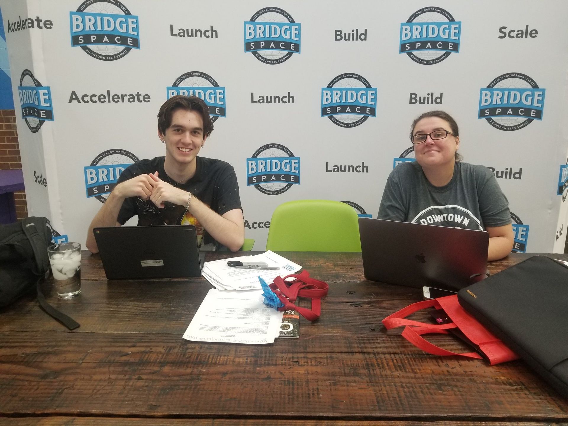 Two people are sitting at a table with laptops in front of a wall that says bridge space