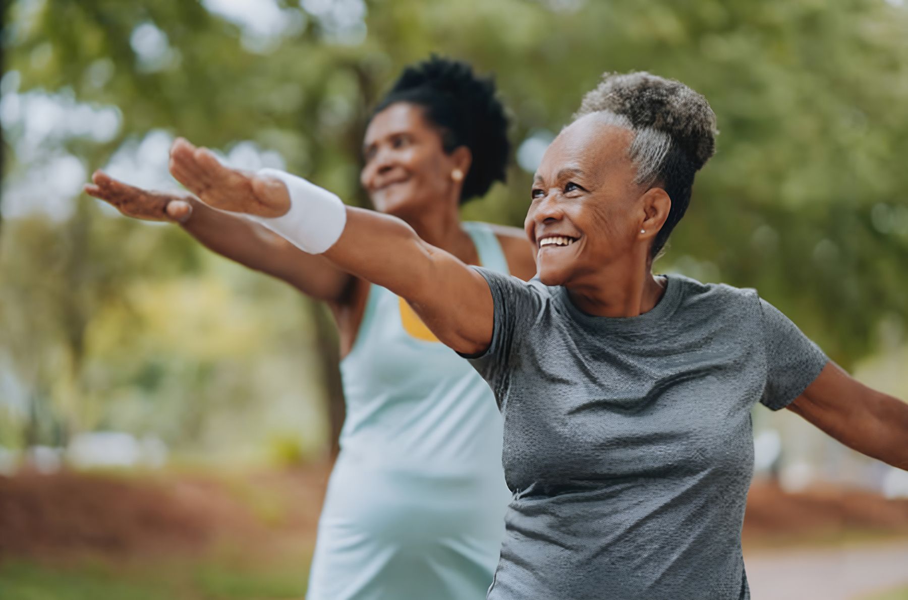 Two smiling older Black women exercising outdoors with arms outstretched in park.