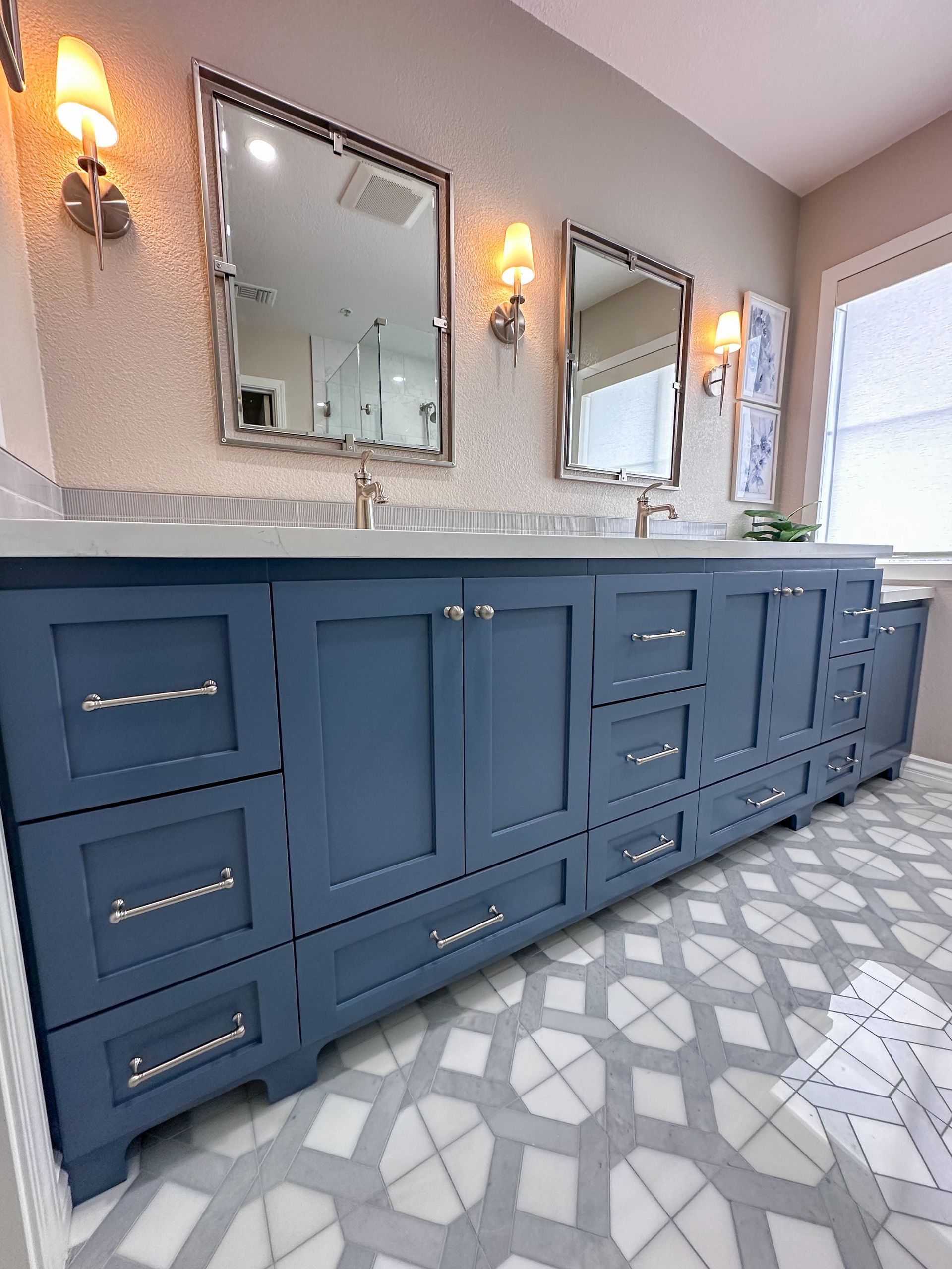 A blue double-sink bathroom vanity with silver hardware, two mirrors, and wall sconces against a patterned floor.