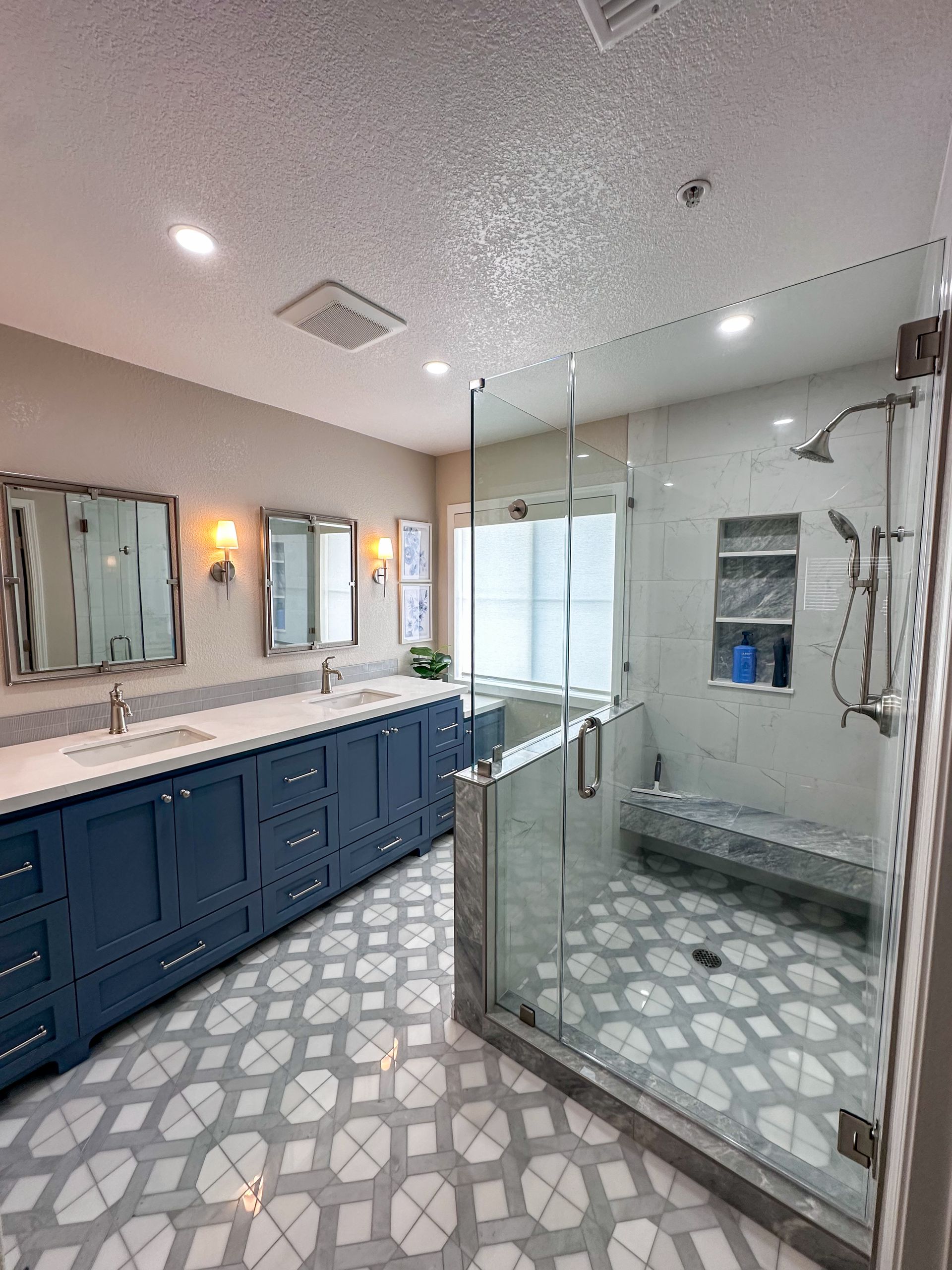 A modern bathroom features a long navy blue double vanity, a glass-enclosed walk-in shower, and patterned floor tiles.